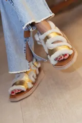 Sandalias de diseño moderno con tiras de cuero metalizado en color dorado, bordeadas con corderito sintético blanco. Poseen una suela plana con plataforma geométrica y cierre con hebilla lateral.