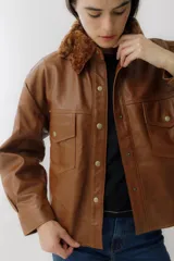 Camisa marrón de cuero sintético con cuello de piel sintética color marrón, cierre frontal con botones a presión dorados, dos bolsillos con solapa en el pecho y corte holgado.