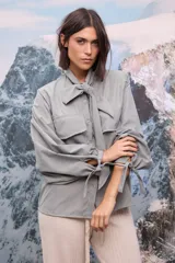 Camisa de tela afelpada color verde grisáceo, con cuello con lazo anudado, mangas largas abullonadas con detalles de lazos en los puños y cierre frontal con botones.