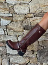 Bota de caña alta estilo biker, confeccionada en cuero color chocolate. Presenta un cierre metálico lateral que recorre toda la caña, estribo y cadena decorativa desmontables, y taco cuadrado con plataforma.