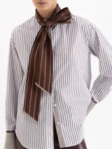 Camisa de poplín con diseño de rayas verticales en tonos azul y blanco. Presenta un cuello prolongado que se anuda en forma de lazo frontal, mangas largas con puños anchos y corte holgado.