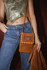 Bandolera pequeña de gamuza color arena, con solapa y detalles de costura en cuero claro. Incluye correa fina de cuero.