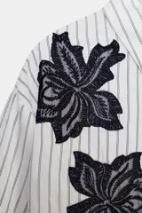 Camisa de cuello solapa y manga larga, con estampado de rayas verticales blancas y grises. Presenta dos aplicaciones de flores de encaje negro en la parte frontal. Cierre frontal con botones ocultos por solapa.