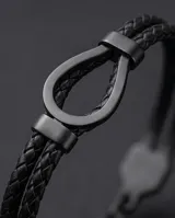 Pulsera tipo esclava compuesta por una doble tira de cuero trenzado negro y un cierre central de acero en forma de herradura.