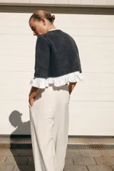 Chaqueta gris de cuello redondo y manga tres cuartos, con detalle de volantes de popelín blanco en el cuello, puños y bajo. Cierre frontal con botones.