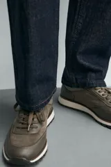 Zapatilla deportiva color verde militar, con parte superior de múltiples piezas que combinan piel y serraje. Presenta horma redonda y suela dentada en color crema con base marrón.