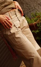 Pantalón ancho de tiro alto color beige, con pliegues frontales y botones visibles en la cintura. El ruedo del pantalón tiene un detalle de terminación en cuero color caramelo.