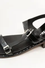 Sandalias negras de cuero con diseño de tiras rectas, hebillas metálicas y un mini taco de 1.5 cm. La suela tiene detalles de tachas pequeñas.