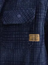Sobrecamisa de tejido polar con estampado abstracto en tonos rojizos y azul marino. Presenta cuello clásico, cierre frontal con botones a presión, dos bolsillos frontales con solapa en el pecho y un parche decorativo.