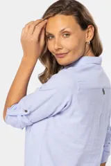 Camisa blanca de manga larga con cuello y tapeta de medio botón. Presenta dos bolsillos frontales con tapa y botones metálicos. Las mangas tienen presillas para sujetar el puño enrollado.