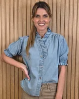 Camisa de denim de algodón celeste, con cierre frontal de botones, cuello alto con volados y mangas cortas abullonadas con puños elastizados fruncidos.