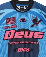 Remera de motocross manga larga marca Deus Ex Machina, en tonos azules con gráficos estampados en color rosa y negro. Presenta cuello redondo, puños elásticos y diseño deportivo con logotipos de la marca.