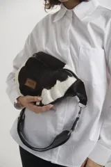 Riñonera de diseño combinado, con frente en cuero vacuno con pelo estampado tipo animal print de vaca en blanco y negro, y cuerpo principal en cuero liso color negro. Cuenta con cierre metálico, correa ajustable y etiqueta de cuero con logo grabado.