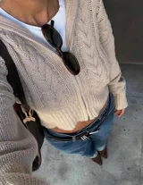 Sweater de punto color beige con diseño de trenzas, cierre frontal completo, cuello alto y mangas largas con puños ajustados. Presenta un corte ligeramente corto.