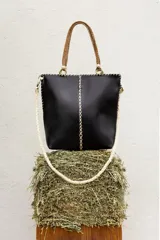 Cartera de cuero negro cosida a mano con costuras visibles decorativas, asas rígidas trenzadas en tonos marrones y correa larga de mano trenzada en color crudo.