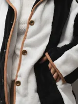 Campera de textura suave y afelpada con diseño de rayas verticales en blanco y negro. Presenta un corte recto, cuello con solapas y cierre frontal con botones. Los bordes están terminados con un ribete en color tostado que aporta contraste y estructura a la prenda.