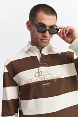 Polo de manga larga con diseño de rayas horizontales anchas en color marrón y crema. Presenta cuello camisero blanco con cartera de botones y estampado frontal con texto decorativo y logo de la marca.