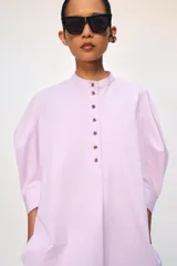 Camisa confeccionada en hilatura de algodón de color rosado claro, con cuello mao y cierre frontal parcial con botones a contraste. Presenta mangas por debajo del codo abullonadas y bajo asimétrico con aberturas laterales.