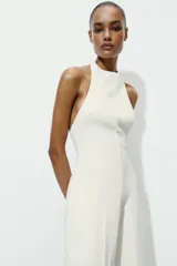 Mono largo de color blanco con cuello halter y espalda descubierta. Presenta un diseño de pierna ancha y cierre posterior con cremallera oculta y botones.