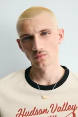 Camiseta de corte regular fit, color crema, con cuello redondo y mangas cortas. Presenta un estampado frontal con un personaje de dibujos animados jugando al baloncesto y texto "Hudson Valley Company Champion '87 NYC, USA". Los acabados del cuello y las mangas son de vivo negro a contraste.