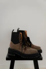Borcego de gamuza color chocolate con paneles laterales elásticos negros y suela track dentada. Presenta sistema de cordones con ganchos metálicos y tirador en la parte trasera.