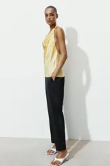 Top de cuello halter con escote fluido y espalda descubierta. Presenta un diseño satinado en color amarillo con estampado de lunares pequeños y un aplique metálico decorativo en el cuello.