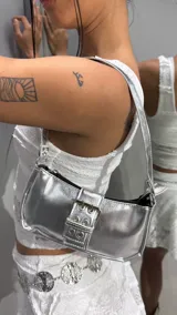 Cartera de hombro plateada con acabado metalizado, correa corta y detalle de tira frontal con hebilla y ojetes metálicos.