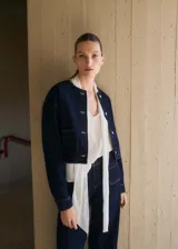 Chaqueta corta de denim color azul oscuro, con cuello redondo, cierre frontal de botones y dos bolsillos de parche delanteros.