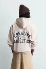 Sudadera hoodie relaxed fit de color beige, con cuello con capucha, manga larga y bolsillo frontal tipo canguro. Presenta un pequeño estampado de texto en contraste en el pecho.