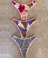 Pack de tres bombachas tipo colaless de algodón acanalado con elástico fino en cintura y contorno de pierna. Incluye una bombacha gris, una con estampado abstracto en tonos marrones y violetas, y otra con estampado abstracto en tonos rosados y marrones.