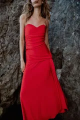 Vestido largo de fiesta color rojo, con escote corazón y diseño palabra de honor. Presenta detalles de drapeado lateral y una superposición de tela con lazada en la cintura, con falda de corte evasé.