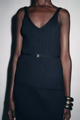 Top de punto acanalado en color azul marino, con escote en pico y tirantes. Presenta un detalle de cinturón fino al tono con un aplique metálico dorado en el centro.