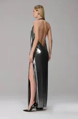 Vestido largo de fiesta en tejido metalizado plateado, con escote profundo tipo degagê y tiras con detalle de cuero en los hombros. Presenta un diseño fluido con tajo lateral pronunciado.