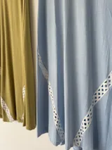 Vestido largo de lino color verde oliva, con cuello redondo, mangas cortas con vuelta y bolsillos laterales. Presenta detalles de pasamanería blanca calada en los laterales de la falda.