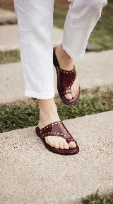 Sandalias chatas de cuero color bordó, con diseño de tira ancha que envuelve el empeine y sujeta el dedo gordo, decoradas con tachas metálicas doradas.