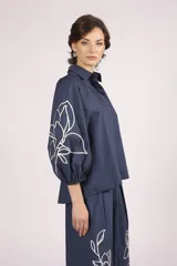 Camisa de algodón color celeste con mangas abullonadas y detalles de bordado lineal blanco en las mangas. Presenta un corte holgado y cierre frontal con botones.