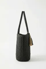 Bolso tipo tote color negro, tejido, con doble asa superior y detalle de borlas colgantes en tonos beige y negro.