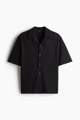 Camisa negra de manga corta con cuello resort, tapeta lisa y bajo recto con aberturas laterales. Corte holgado.