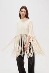 Poncho de punto color crudo con cuello alto y diseño de flecos largos en el ruedo.