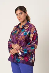 Blusa tipo casaca italiana abotonada, de satén estampado con fondo violeta y flores en colores azul, mostaza y naranja. Tiene cuello de camisa y mangas largas con presilla para sujetar.
