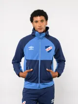 Campera deportiva con capucha, confeccionada en tejido técnico de nylon con bloques de color en azul marino y celeste. Presenta cierre frontal, bolsillos laterales y el escudo del Club Nacional de Football junto al logo de Umbro en el pecho.