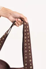 Cartera tipo tote de gamuza color tostado, con diseño triangular y correas integradas decoradas con tachas metálicas doradas a lo largo de los bordes.