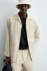 Sobrecamisa de hombre de corte relajado, color beige claro con finas rayas verticales en tono más oscuro. Presenta cuello bowling, manga larga con puño abotonado, bolsillos de parche en el pecho y en la cadera, y aberturas laterales en el bajo.