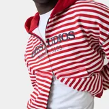 Campera con capucha de felpa de algodón, con diseño a rayas horizontales rojas y blancas, cierre frontal con cremallera bidireccional y bolsillos tipo canguro. Presenta grandes letras bordadas con el texto "EMESTUDIOS Madrid, 5135" en el pecho.