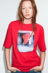 Camiseta de algodón color rojo, corte regular fit, cuello redondo y manga corta. Presenta una estampa fotográfica en el centro con dos personas sosteniendo un gran corazón rojo inflable. Incluye texto con nombre y copyright.
