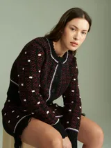 Cardigan de punto negro y rojo con diseño texturizado, cuello redondo y cierre frontal con botones. Presenta detalles de ribetes en blanco y violeta en el cuello, la botontera, los puños y el ruedo, que cuenta con flecos colgantes.