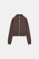 Chaqueta de felpa color marrón chocolate, con cuello solapa, cierre frontal con cremallera y bolsillos de plastrón en el delantero. Presenta mangas largas con puños ajustados.