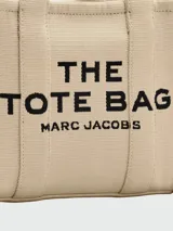 Cartera tipo tote bag de lona color beige, con diseño estructurado y asas superiores. Presenta el logo de la marca estampado en la parte frontal y detalles de cintas laterales con inscripciones.