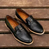 Mocasín de cuero vacuno negro con textura grabada tipo reptil. Presenta un diseño clásico con antifaz en el empeine, suela de goma en tono natural y taco bajo de madera.