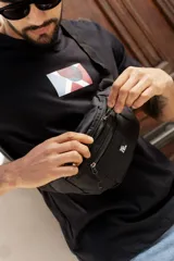 Riñonera negra de diseño funcional con dos compartimentos principales con cierre y correa ajustable. Presenta una etiqueta rectangular con logo en el frente.